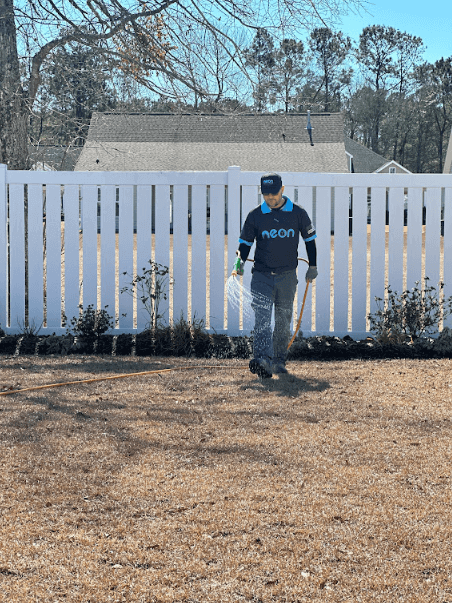 A Neon Pest and Lawn employee watering a client's yard, with a white fence in the background.