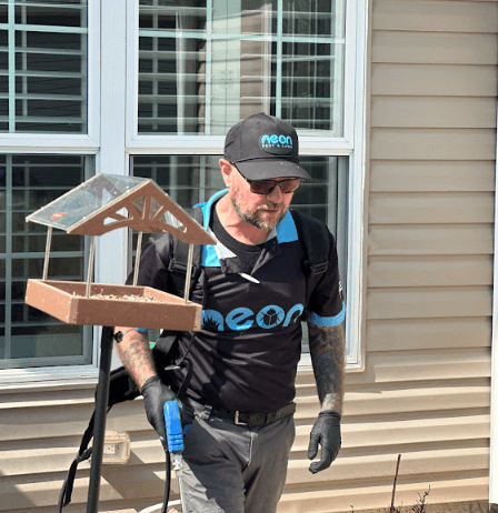 A man in a black shirt and hat carries a bird feeder, representing Neon Pest and Lawn's commercial pest control service.