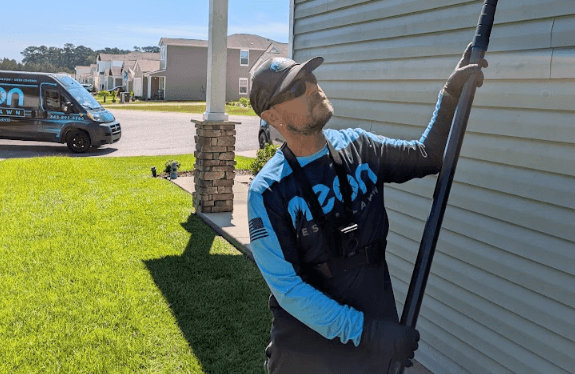 A man in a blue shirt and black hat from Neon Pest and Lawn holds a pole while providing ant control service at a client's home.