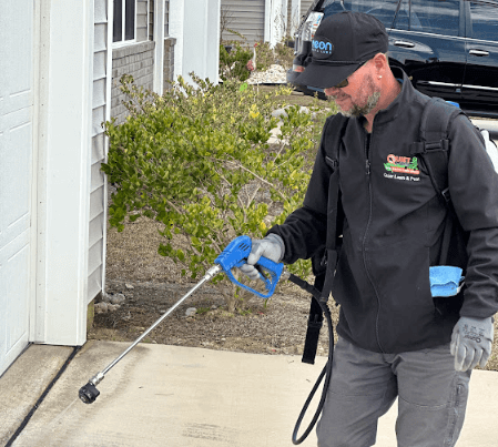A man in a black jacket and hat uses a sprayer on a driveway, providing roach control service for Neon Pest and Lawn.